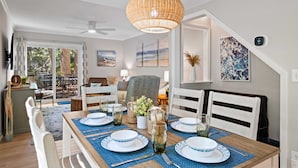 A dining area with a wooden table set for four, surrounded by white chairs. There is a large woven pendant light above the table, and artwork is displayed on the walls.