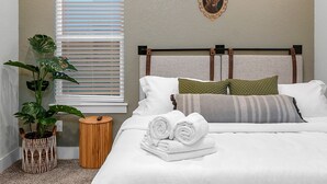 Ein ordentlich bezogenes Bett mit weißen Leinentüchern und aufgerollten Handtüchern, eine Zimmerpflanze, ein kleiner Nachttisch aus Holz und ein Fenster mit Jalousien.