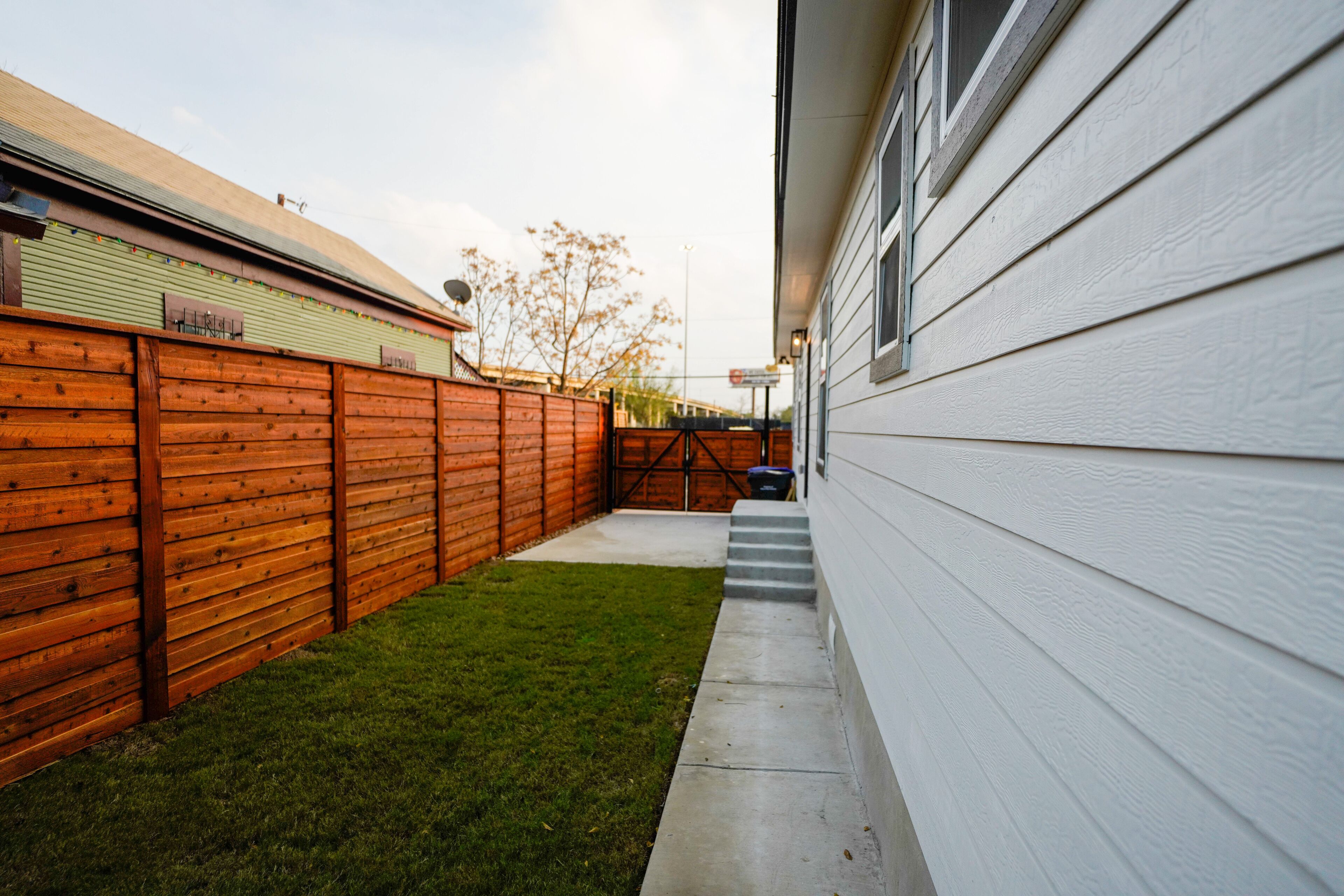 Un jardin avec une clôture en bois, un chemin en béton et un bâtiment blanc.