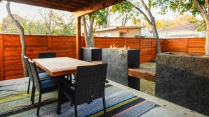 Une terrasse extérieure avec une table et des chaises en bois, un espace barbecue en pierre et une clôture en bois.