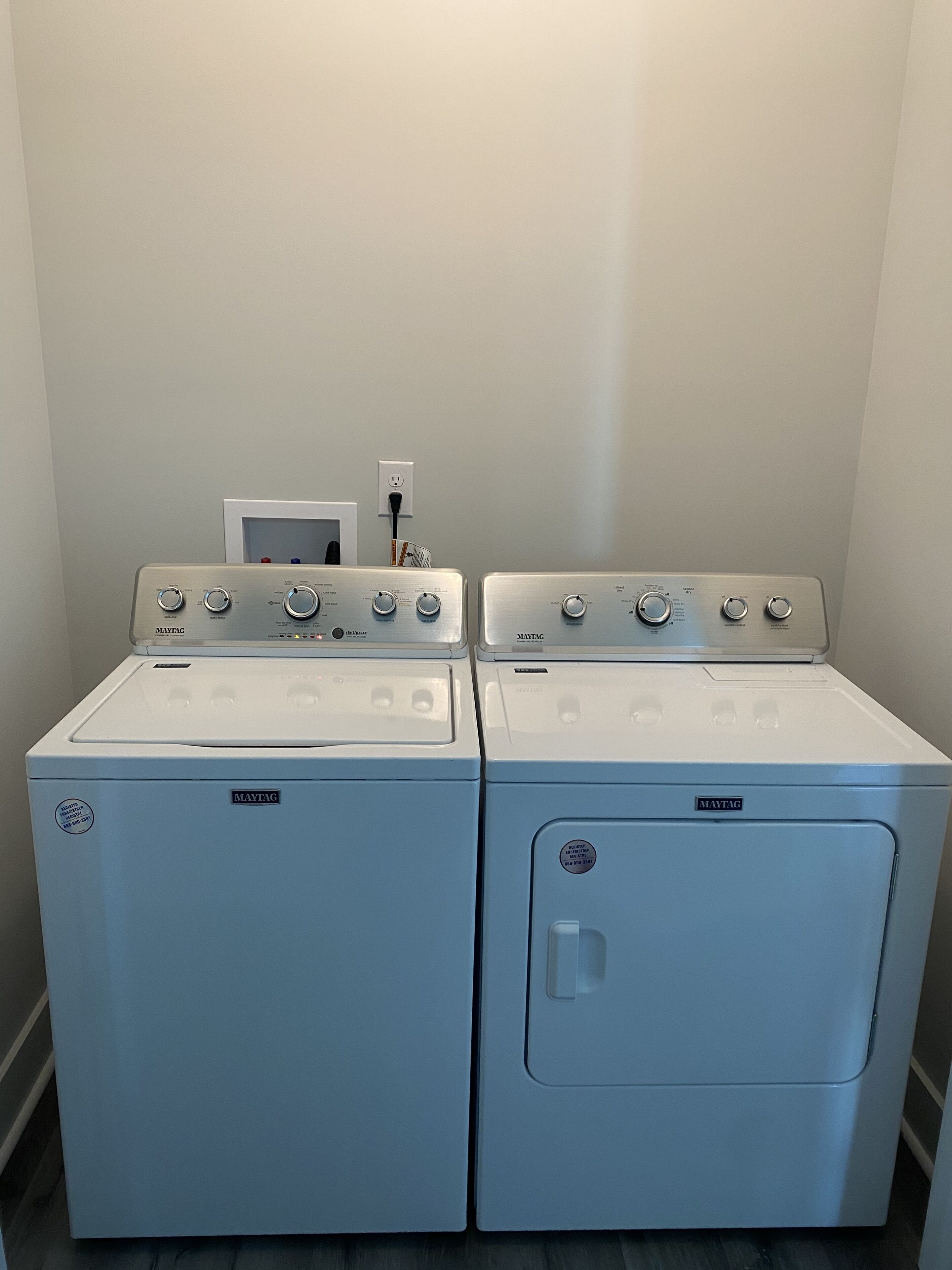 Dos lavadoras y secadoras Maytag blancas de carga frontal una al lado de la otra en un cuarto de lavado con pared blanca y una ventana pequeña.