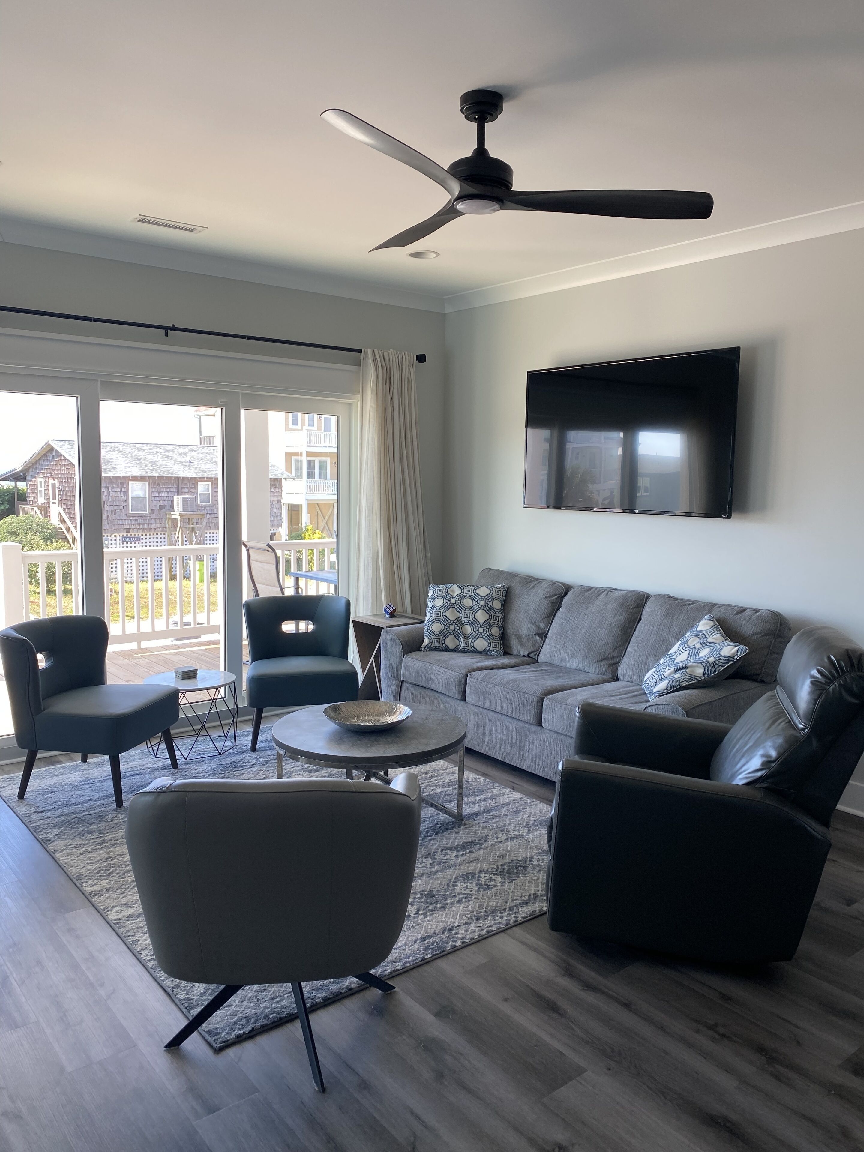 A modern living room with a ceiling fan, a grey sofa, two armchairs, a coffee table, and a TV mounted on the wall.