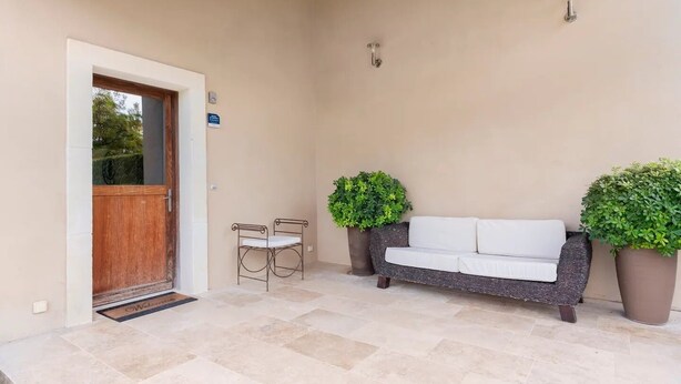 Un espace extérieur aménagé, abrité, avec un canapé et deux fauteuils, une porte en bois et deux plantes en pot.