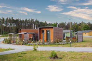 A row of modern, wooden cabins with a central door, surrounded by greenery and a paved path. - Modern Swedish house Lotta #4 on Lake Bärwald - SKAN-PARK (Klitten)