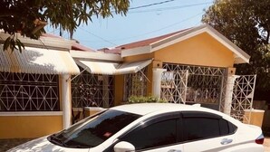 Un coche blanco estacionado frente a un edificio amarillo con entrada protegida por una verja y un porche cubierto.