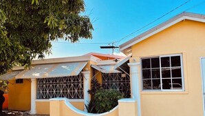Un edificio amarillo con un porche cubierto, una ventana con rejas y un árbol de fondo.