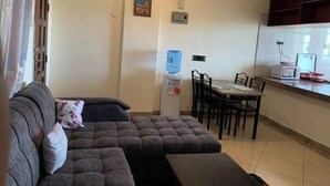 Ein Wohnzimmer mit grauem Sofa, einem Esstisch mit Stühlen, einem Wasserspender und einer Küchenzeile mit Mikrowelle und Toaster.