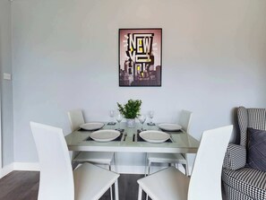 Un coin repas avec une table dressée pour quatre personnes, des chaises blanches et une œuvre d’art représentant un paysage urbain accrochée au mur.