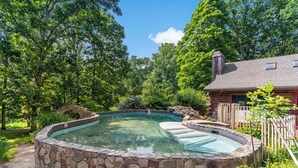 A stone-lined hot tub surrounded by a wooden deck, set in a lush garden with trees and a house in the background. - Guilford Carriage House at Granite Lodge! (Guilford)