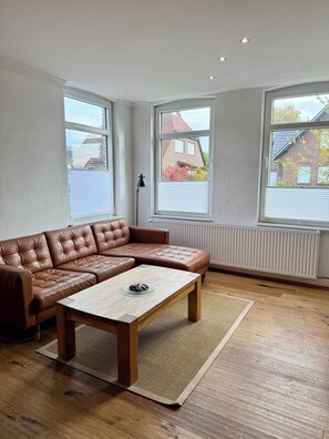 A modern living room with a brown leather sectional sofa, a wooden coffee table, and a large window with a view of a brick house.
