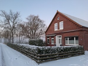 Ein rotes Backhaus mit weißer Tür und Fenstern, umgeben von schneebedeckten Hecken und Bäumen.