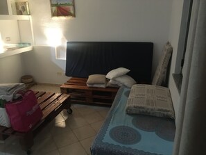 Quarto pequeno e acolhedor com uma cama, um banco de madeira e uma mesinha. Uma janela oferece vista para a paisagem.