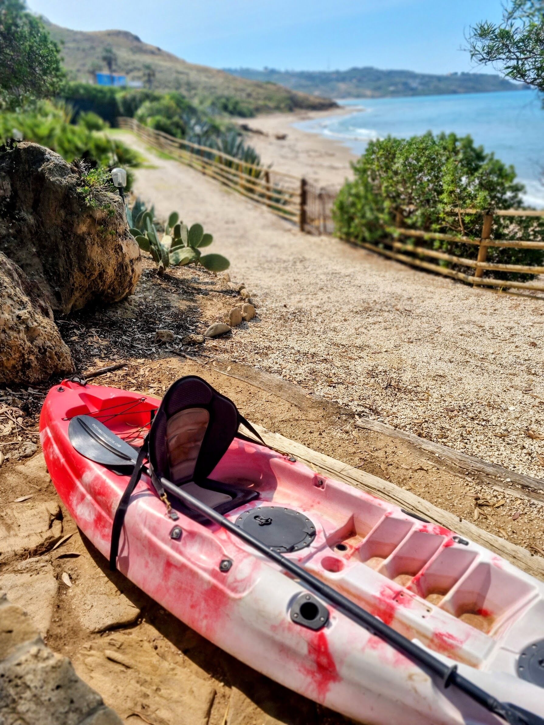 A red kayak with a paddle and a seat on a sandy beach with a wooden fence and greenery in the background.
