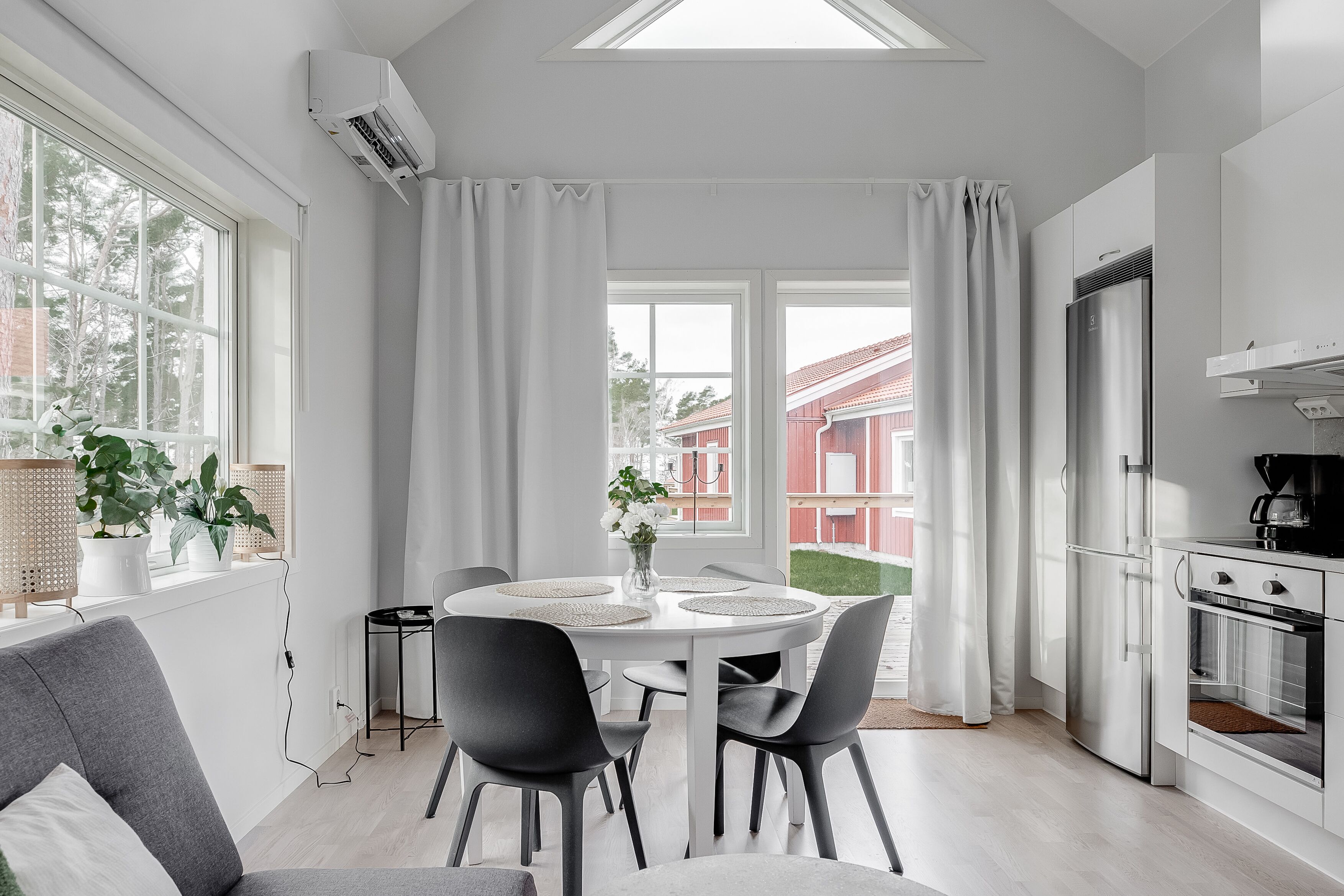 Une cuisine moderne dotée d’un four et d’un réfrigérateur encastrés, un coin repas avec une table et des chaises blanches, et de grandes fenêtres ornées de rideaux blancs.