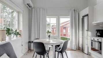 Une cuisine moderne dotée d’un four et d’un réfrigérateur encastrés, un coin repas avec une table et des chaises blanches, et de grandes fenêtres ornées de rideaux blancs.