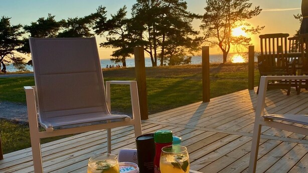 A wooden deck with a table set for two, featuring a bowl of watermelon slices, two glasses of a yellow beverage, condiments, and a smartphone. - Stora Frögården (Morbylanga)
