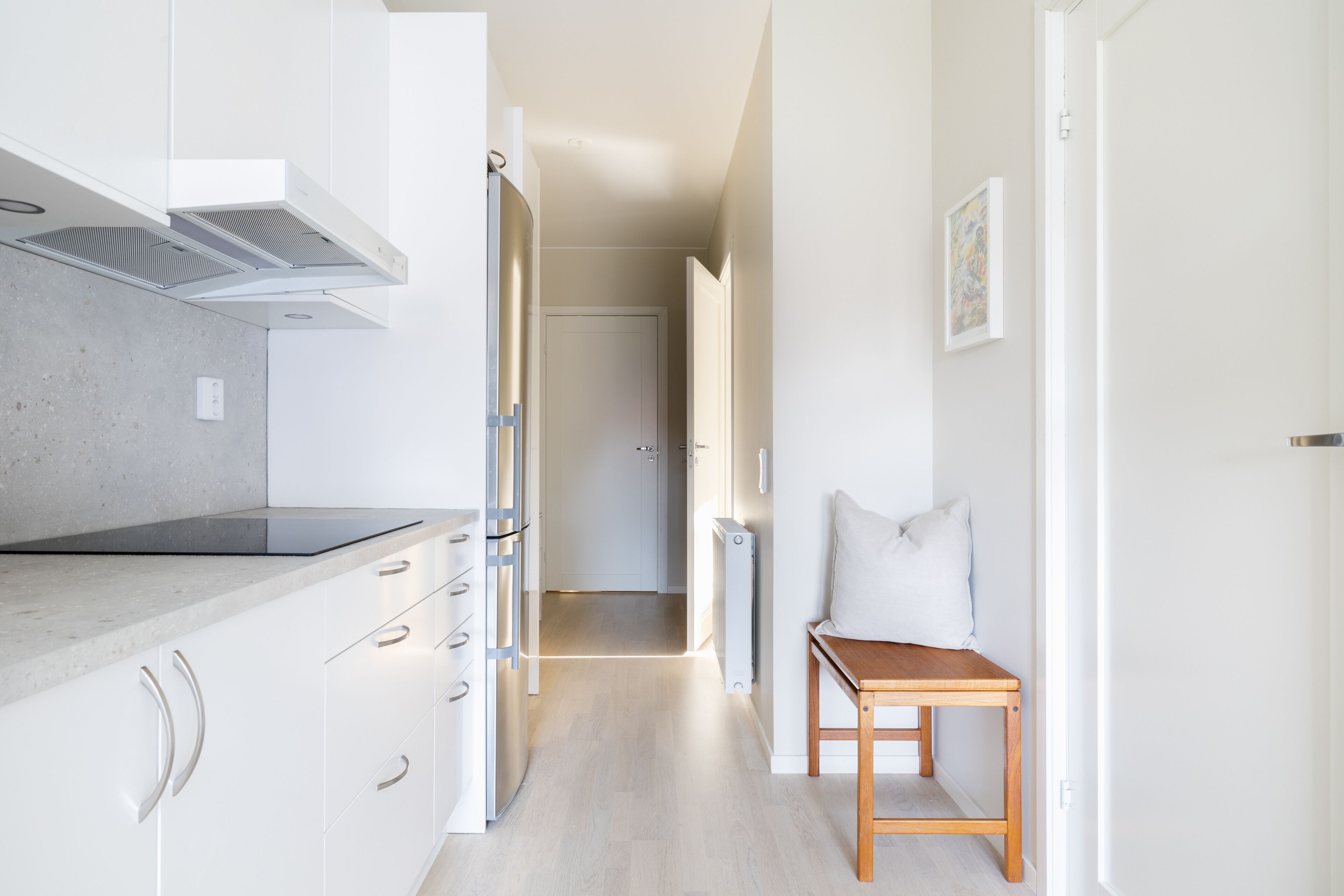 Un couloir étroit, avec un coin cuisine d’un côté, comprenant des meubles blancs, un plan de travail et une cuisinière encastrée. Un banc en bois avec des coussins est placé près de l’extrémité du couloir.