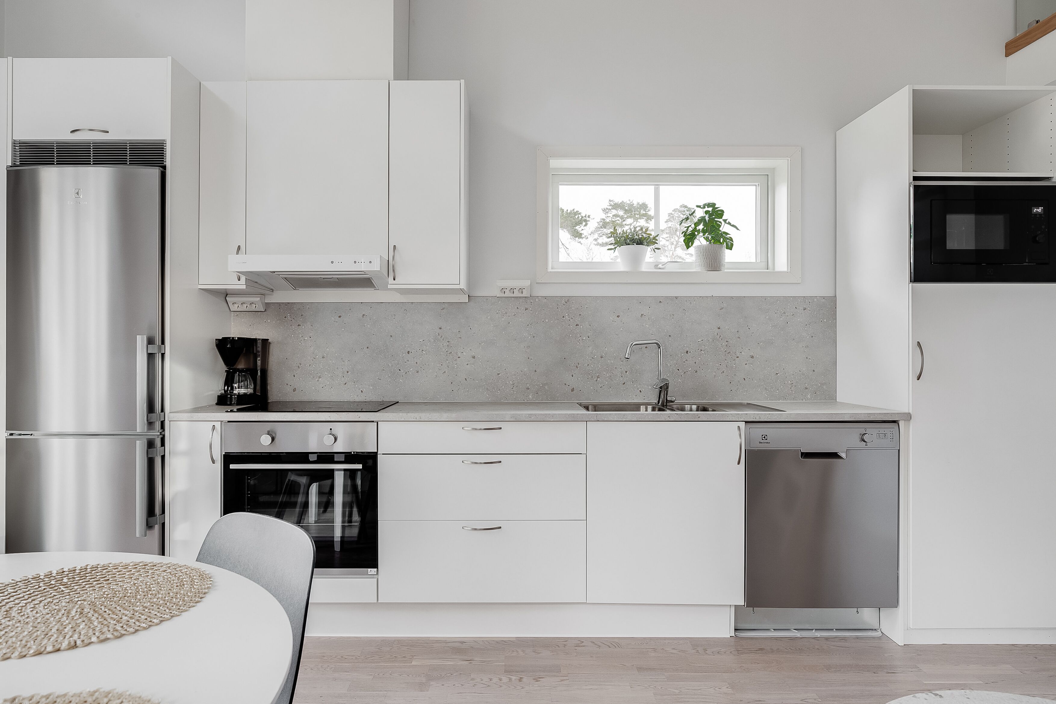 A modern kitchen with white cabinetry, a built-in oven, a dishwasher, and a window with plants.