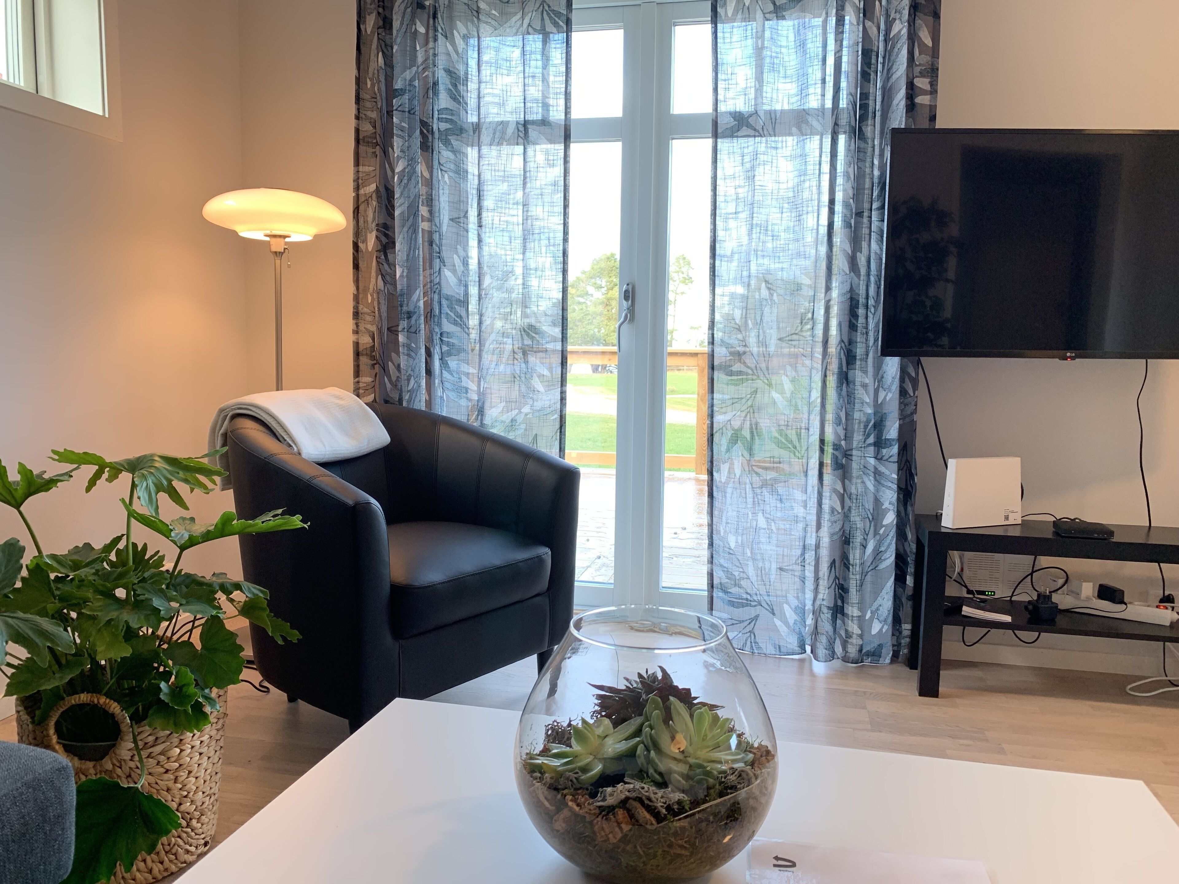 A living room with a black leather chair, a glass table with a succulent plant, a flat-screen TV, and a large window with curtains.