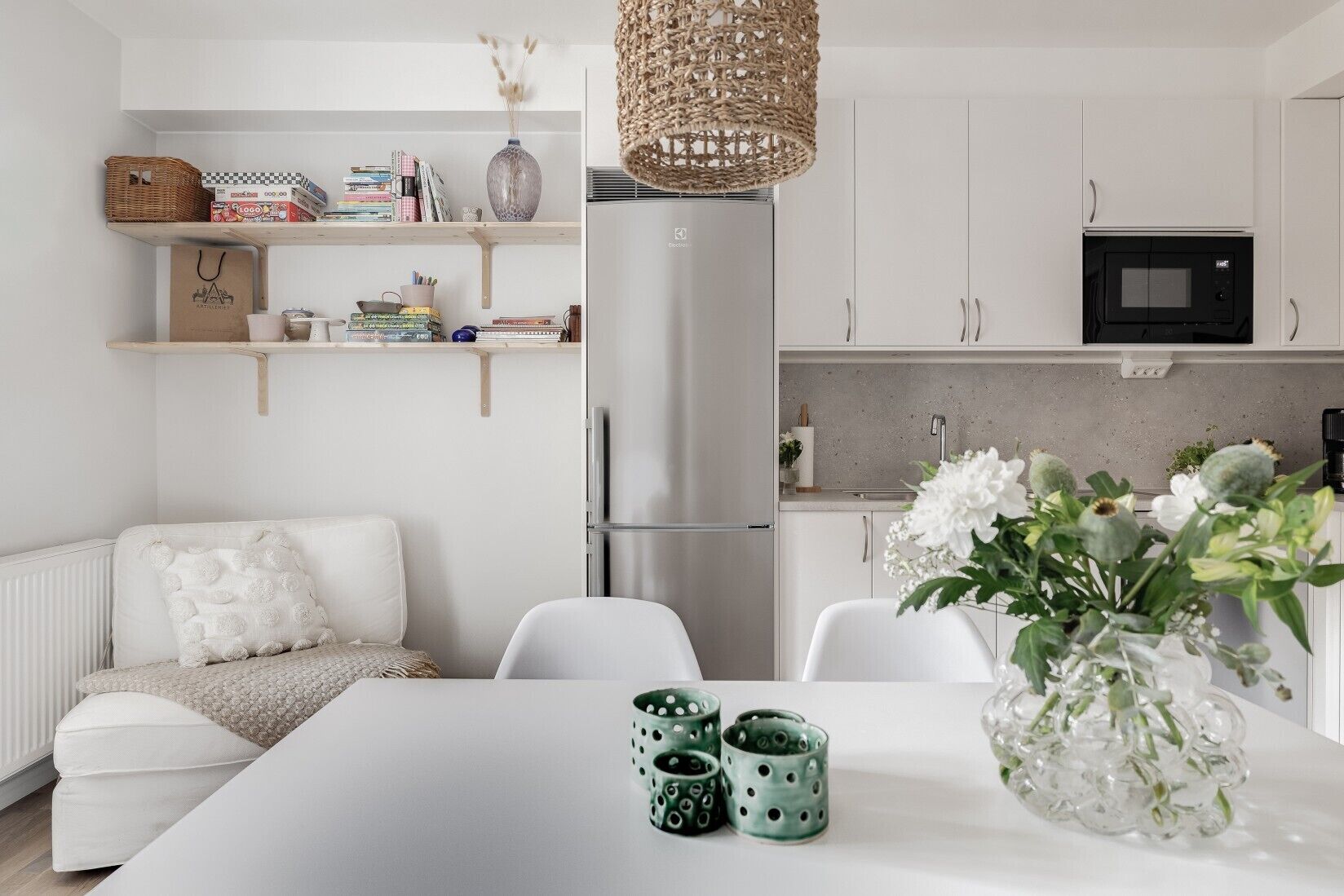 A modern kitchen with white cabinetry, a stainless steel refrigerator, and a microwave. There is a dining table with a vase of flowers and two green mugs.