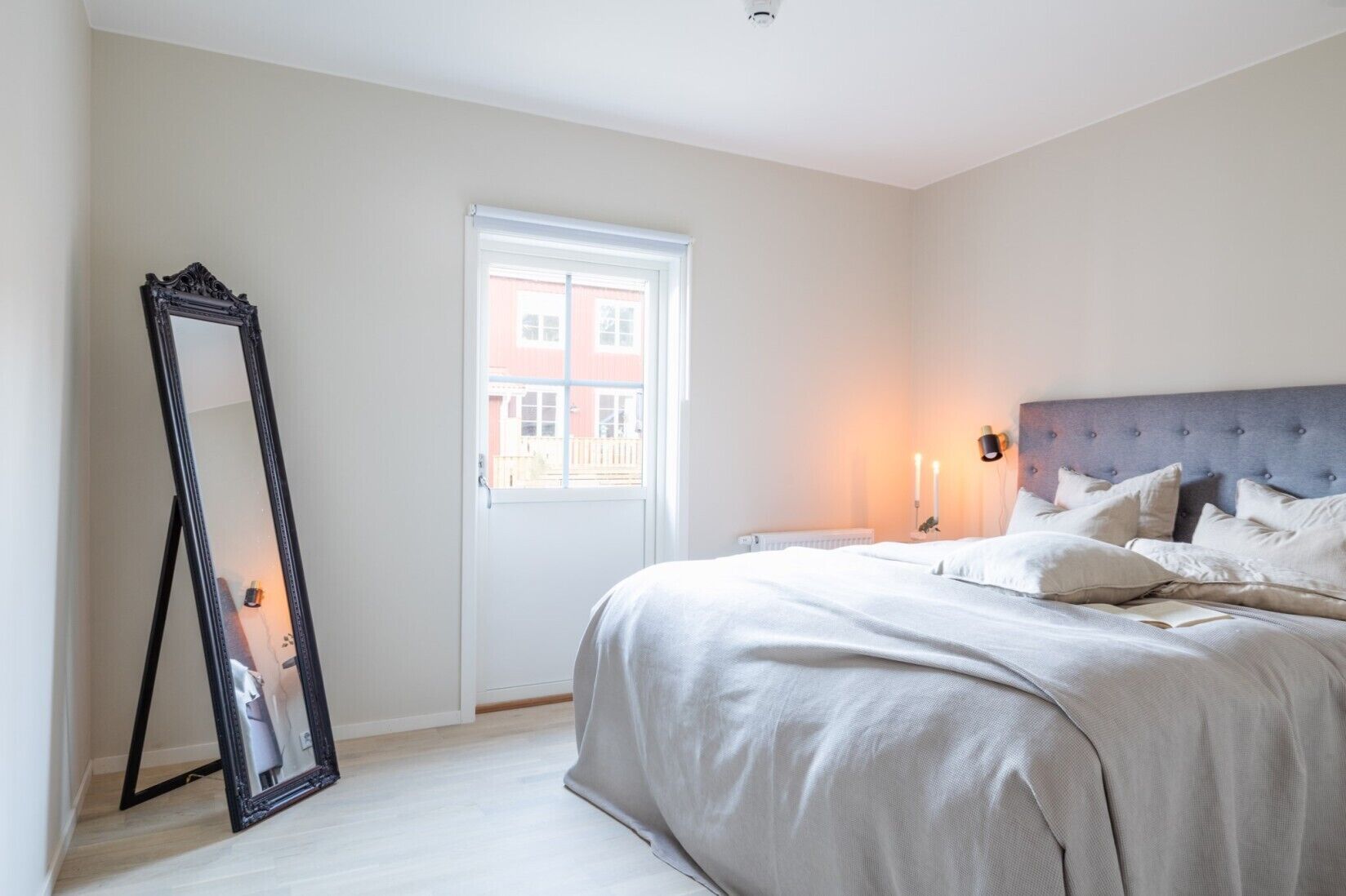 Een slaapkamer met een bed, een spiegel, een raam en een wandlamp.