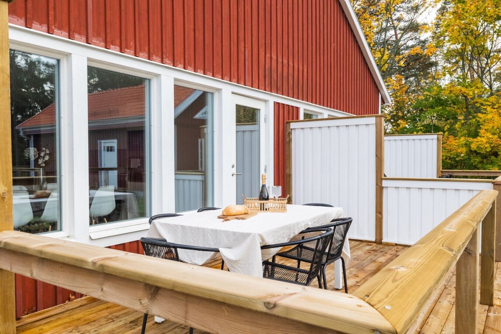 Una terraza de madera con una mesa lista para comer, ventanas amplias y un edificio rojo al fondo.