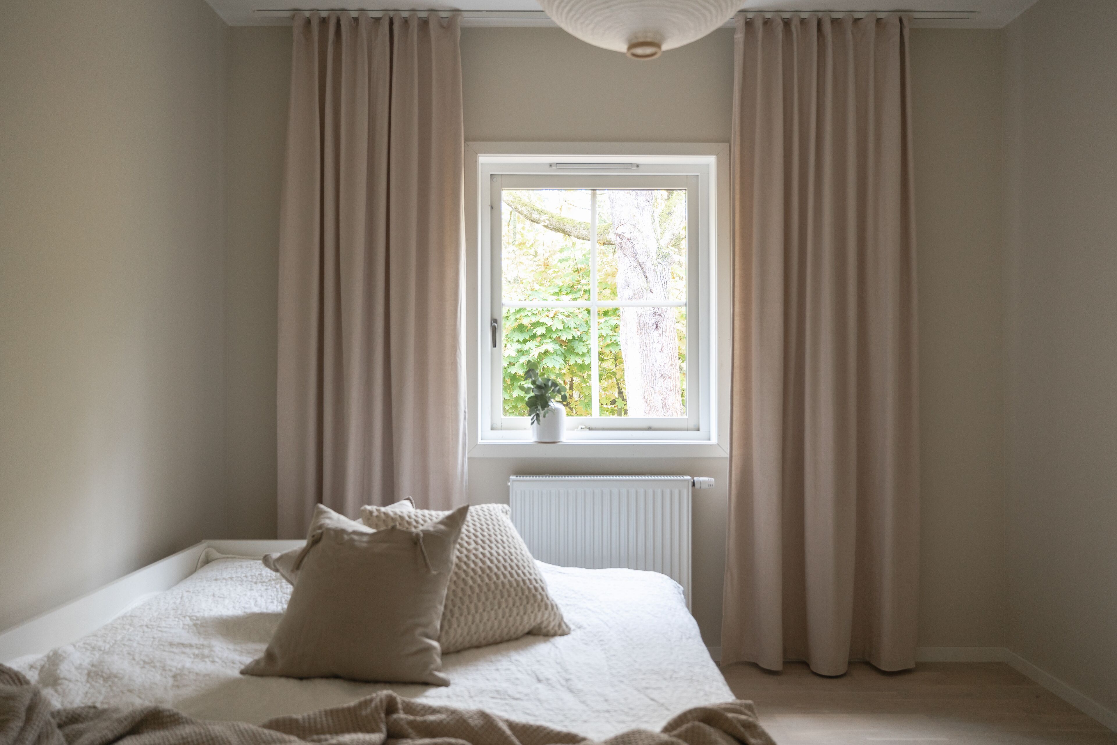 A bedroom with a large bed, beige curtains, a window with a view of greenery, and a radiator.