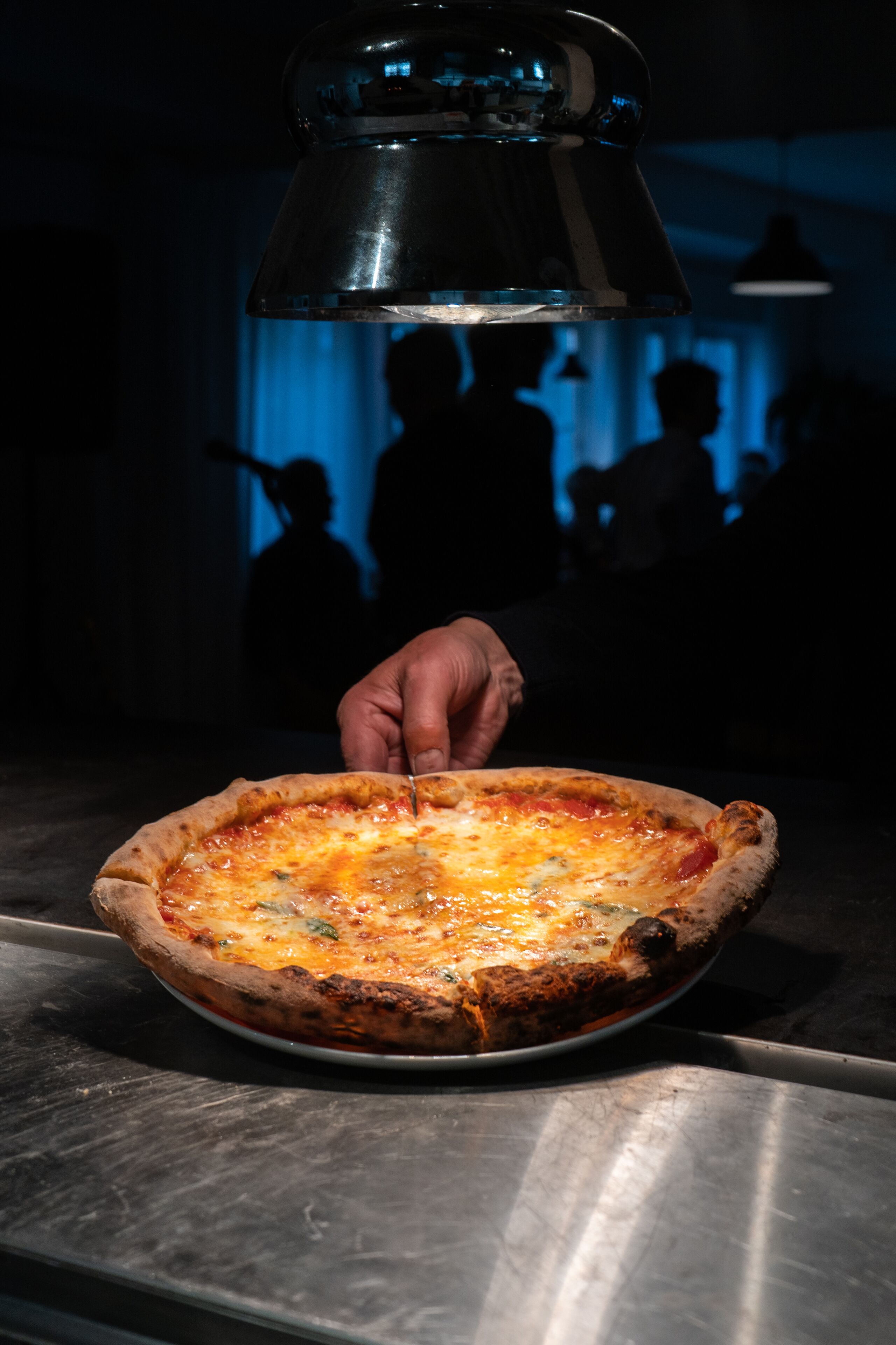 A pizza on a metal tray with a hand holding a knife, set against a dark background with silhouettes of people.