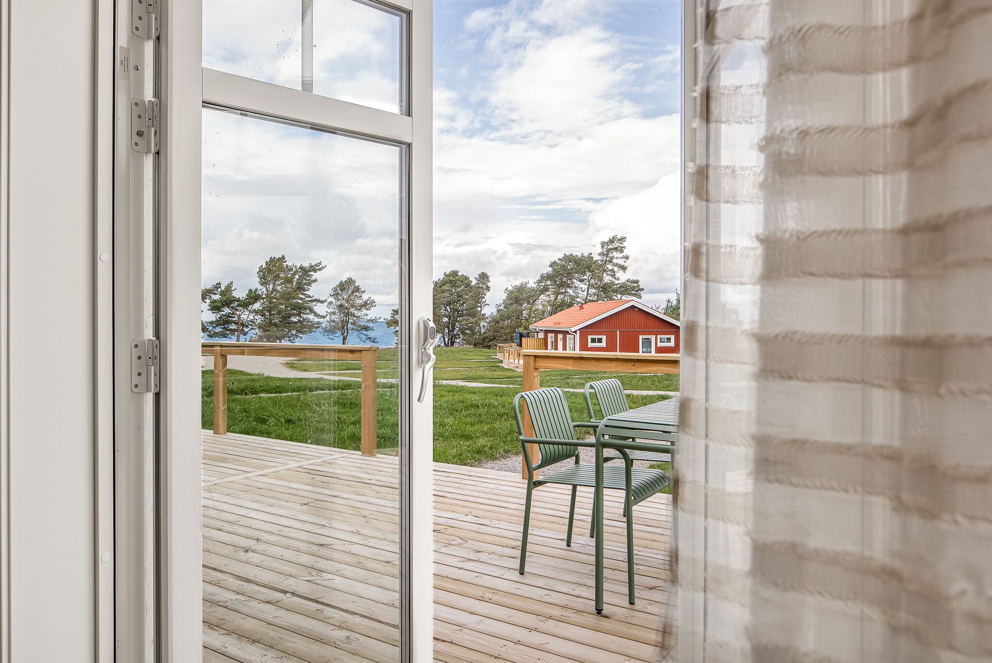 Uitzicht vanuit een kamer met een houten terras, een tafel met stoelen, en een gebouw met een rood dak op de achtergrond.