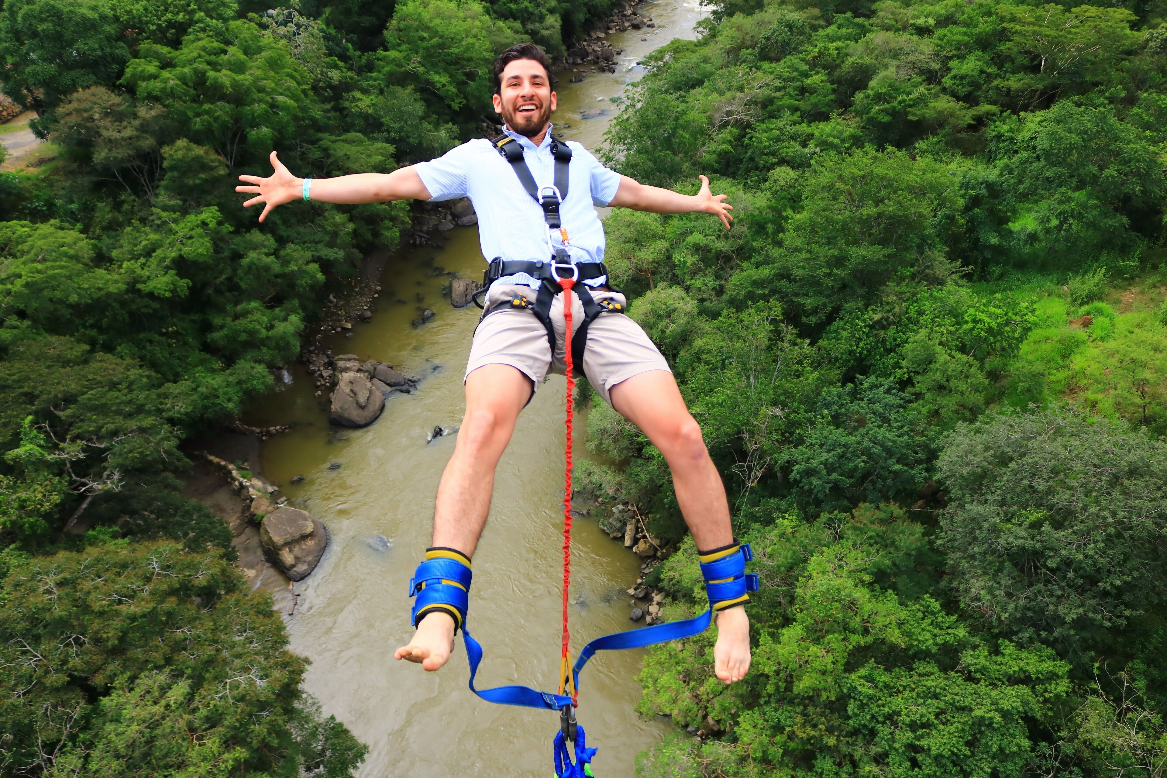 A person bungee jumping with outstretched arms, surrounded by dense green foliage and a river below.