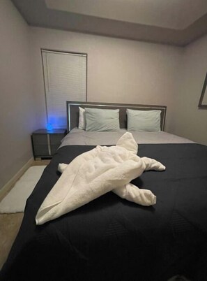 Ein ordentlich bezogenes Bett mit grau-weißem Überwurf, ein Nachttisch mit Lampe und ein Fenster mit Jalousien.