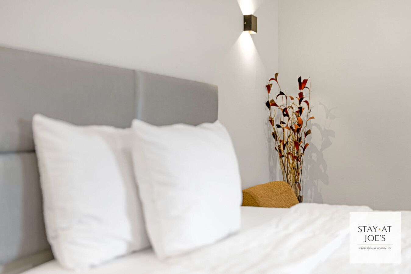 Un lit bien fait, avec une literie blanche et une tête de lit grise, un petit vase décoratif contenant des fleurs séchées et un luminaire fixé au mur.