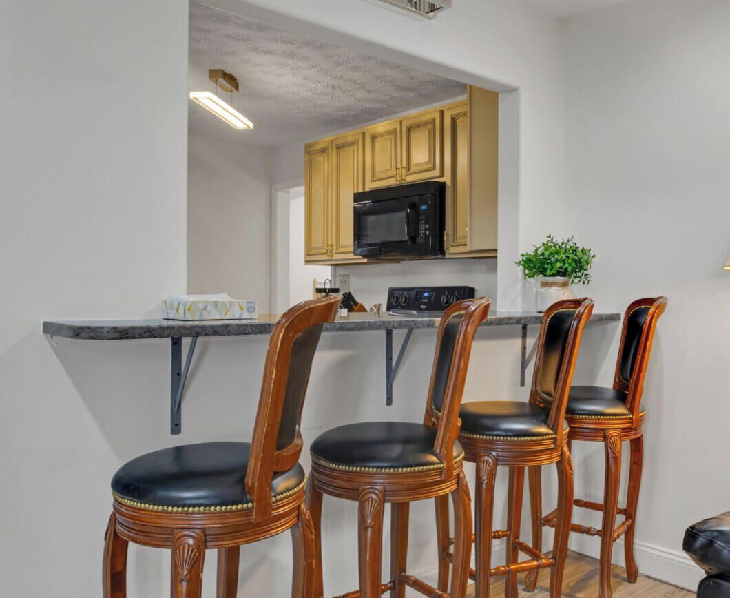 A kitchen with a microwave, a coffee machine, and a plant on the counter. There are three wooden chairs with black cushions.