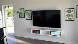 A modern living room with a flat-screen TV mounted on the wall, a built-in shelf below it, and framed artwork on the walls. - Blue Hole Lodge - Bonefish Heaven with Blue Hole in Backyard (Exuma)