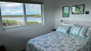 A bedroom with a large window overlooking a body of water and mangroves, a bed with striped bedding, and two framed artworks on the wall. - Blue Hole Lodge - Bonefish Heaven with Blue Hole in Backyard (Exuma)