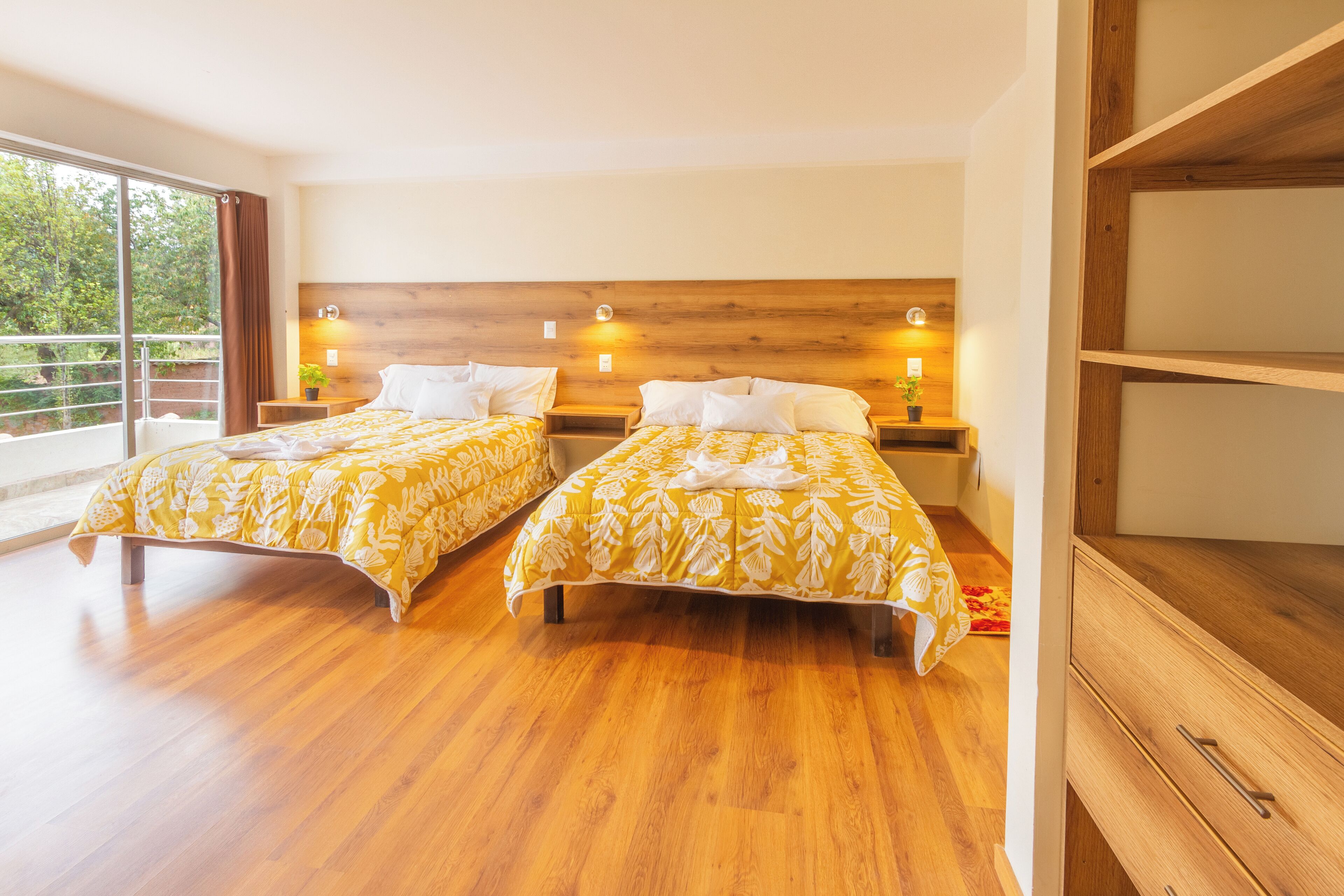 A hotel room with two beds, wooden headboard, bedside lamps, and a large window with a view of greenery.
