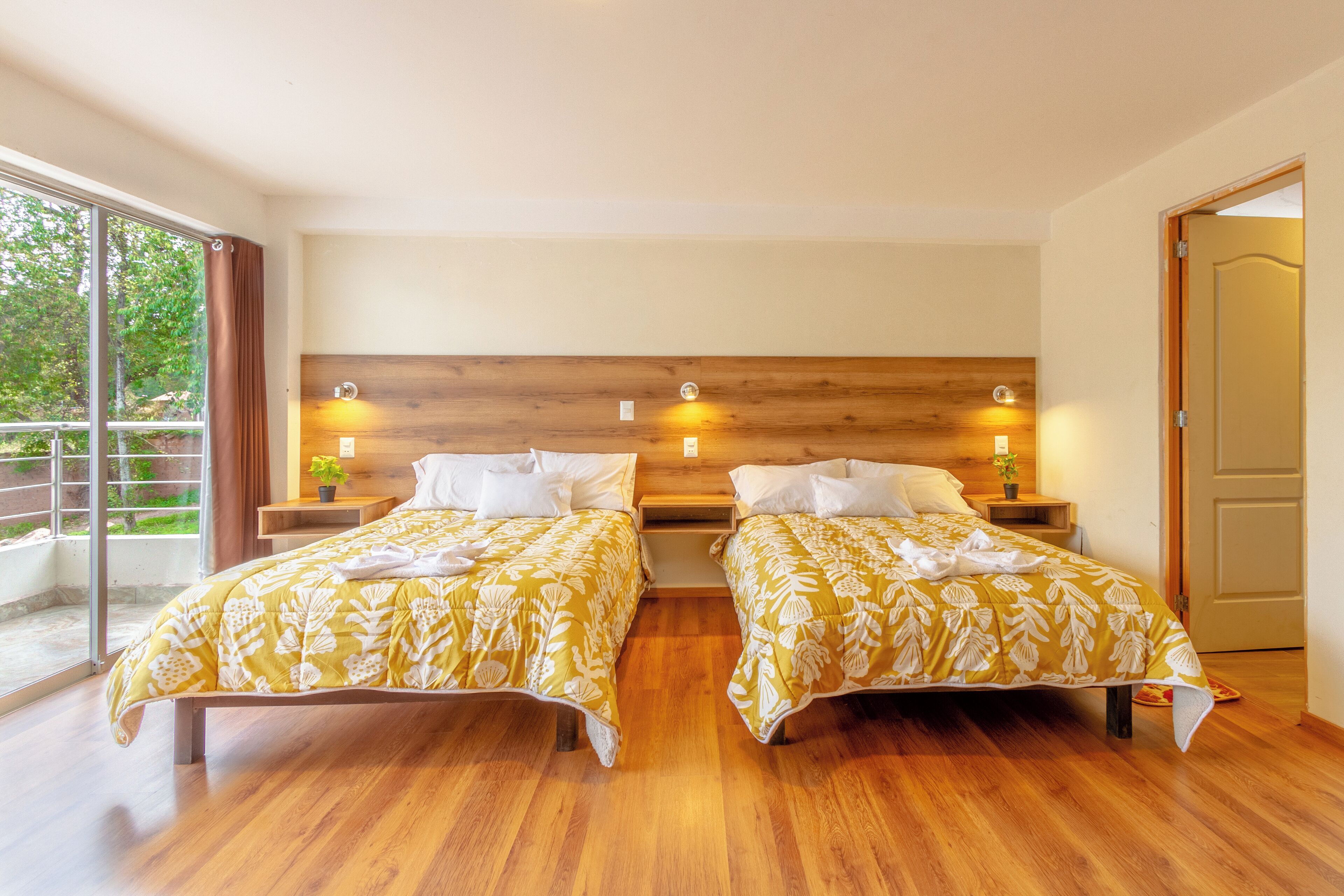 Habitación doble con dos camas, cabecero de madera, mesitas de noche con lámparas y un amplio ventanal corredero a un balcón.