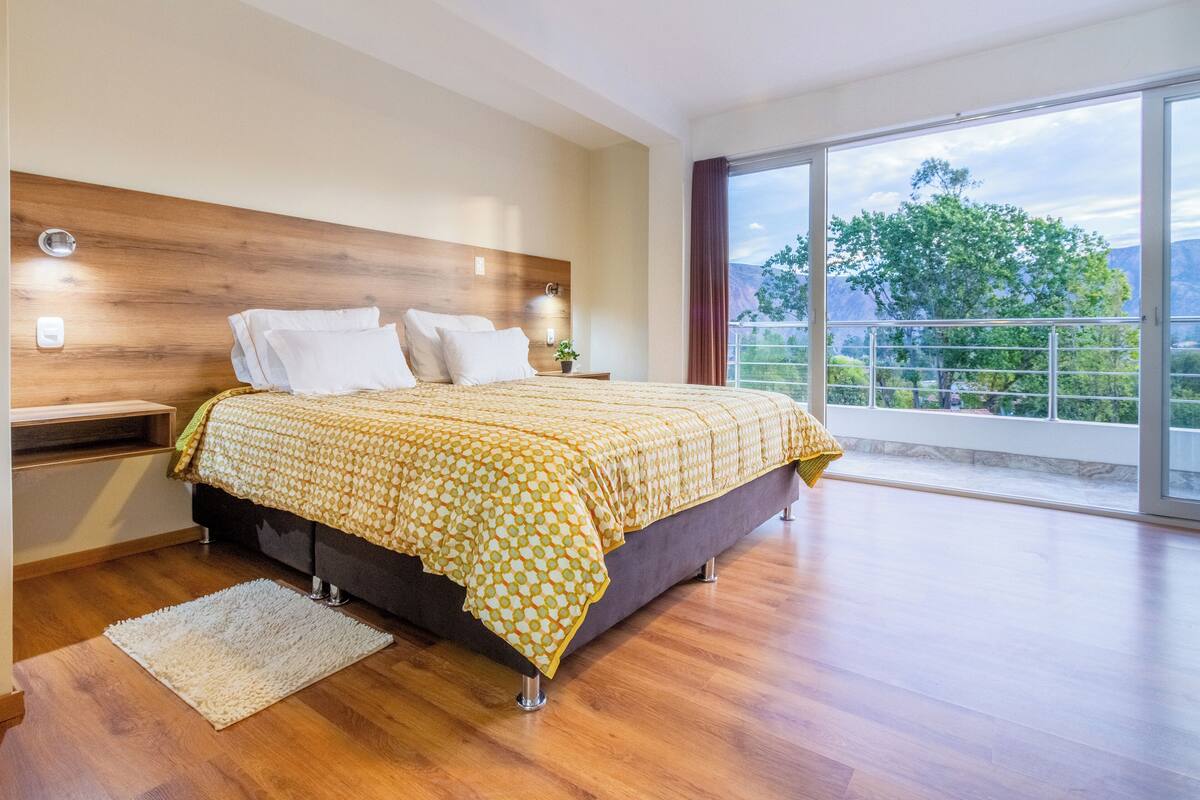 Ein modernes Schlafzimmer mit einem hölzernen Kopfteil, einem großen Bett mit gemustertem Bettbezug, einem Nachttisch mit Lampe und Blick auf Grünflächen und Berge durch große Fenster.