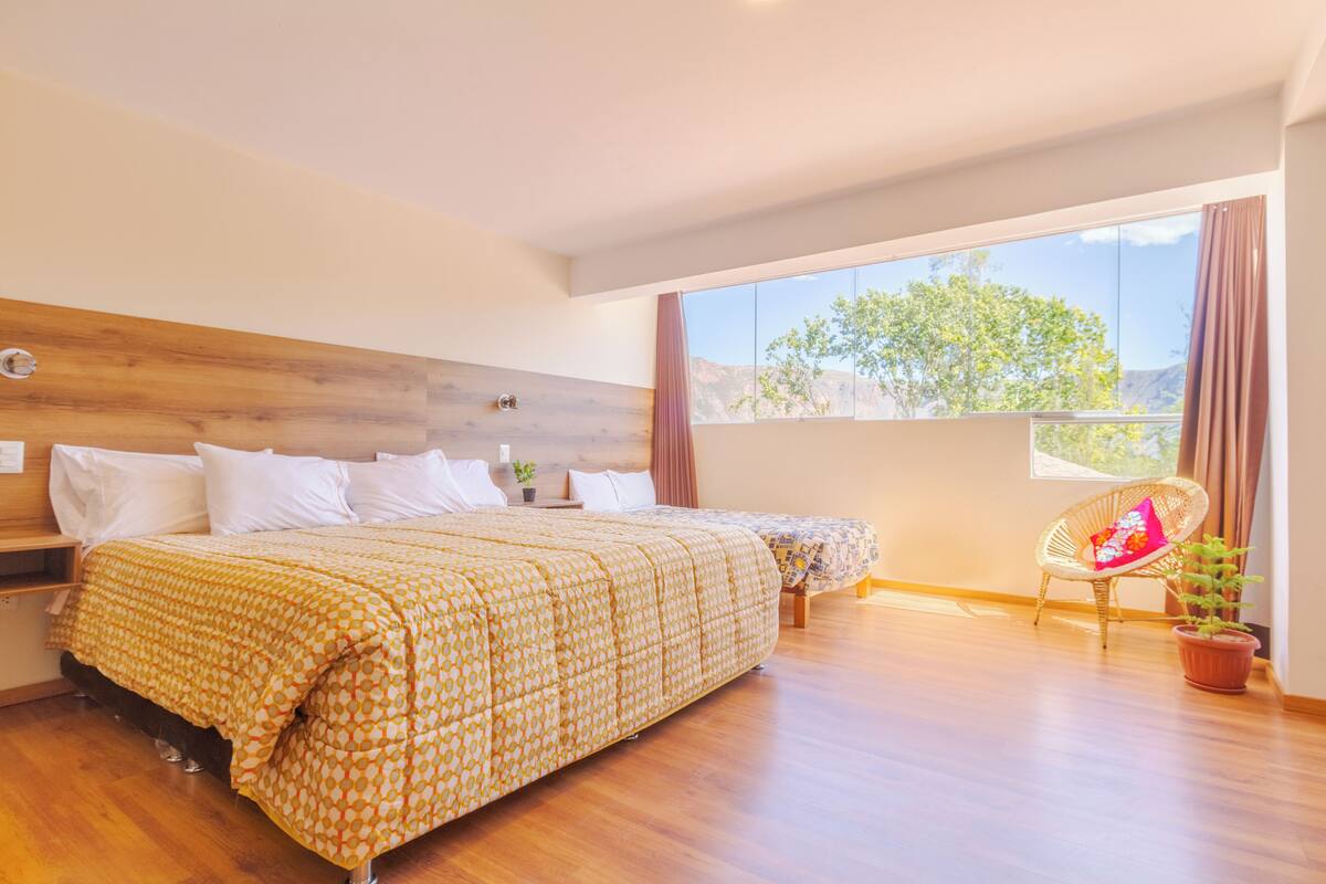 Ein Schlafzimmer mit einem großen Bett, einem Nachttisch, einem Sessel und einem Fenster mit Blick auf Bäume und Berge.