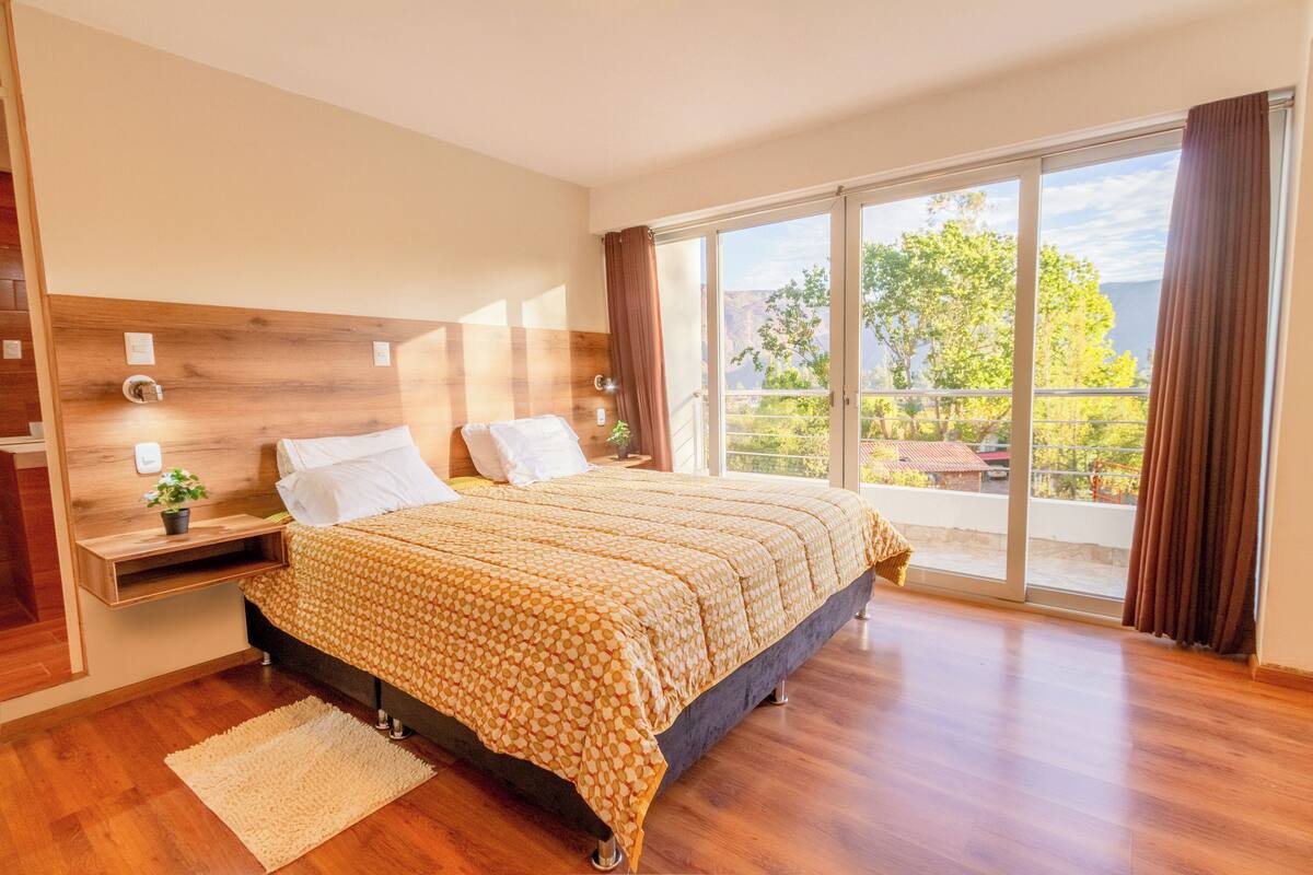 Ein geräumiges Schlafzimmer mit einem großen Bett, einem Holzkopfteil, einem Nachttisch mit einer Pflanze und Blick ins Freie durch eine Schiebetür.