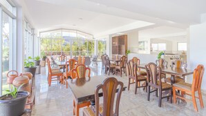 A spacious dining area with high ceilings and large windows, featuring wooden tables and chairs, potted plants, and a kitchen in the background. - Valles apartamentos ecológicos (Urubamba)