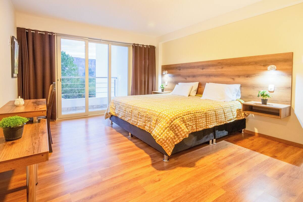 Ein Schlafzimmer mit einem großen Bett, einem hölzernen Kopfteil, einem Nachttisch mit Lampe, einem Schreibtisch mit Stuhl und Blick ins Freie durch eine Schiebetür.