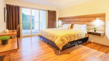 Un dormitorio con una cama grande, cabecera de madera, una mesita de noche con lámpara, un escritorio con silla y vistas al exterior a través de una puerta corredera.