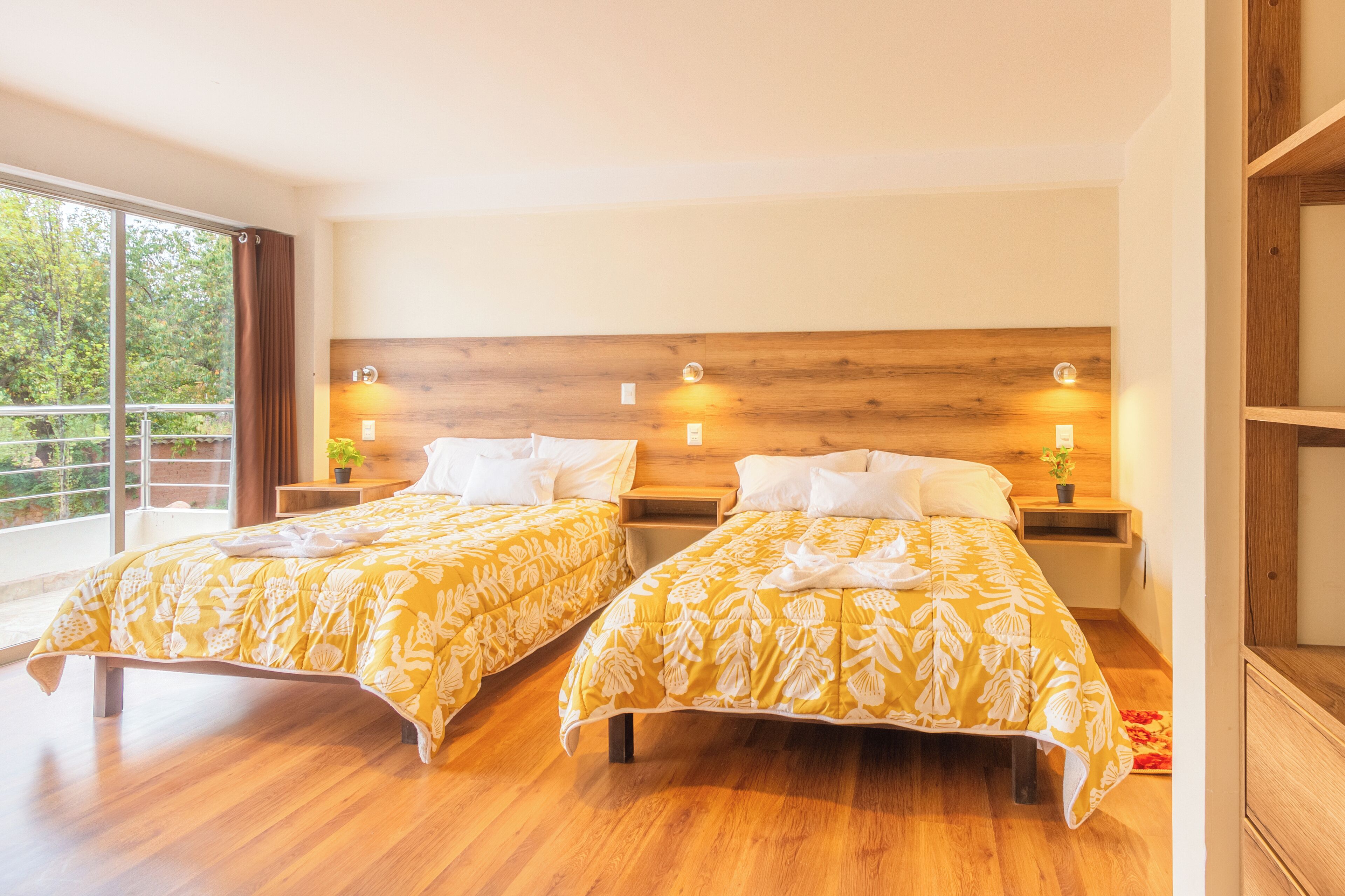 Habitación de hotel con dos camas, cabeceros de madera, mesitas de noche y un amplio ventanal con vistas a zonas verdes.