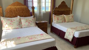 Cama doble con ropa de cama floral y cabecera de madera, una mesita de noche y una ventana con cortinas.