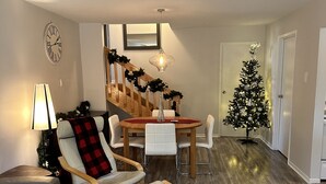 Uma sala de estar com uma mesa de jantar de madeira, cadeiras brancas, um sofá cinzento e um pinheiro de Natal.