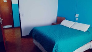 A room with a bed, a nightstand, a door, and a wall with a light switch. - HOTEL LOS ANTIGUOS (Los Antiguos)