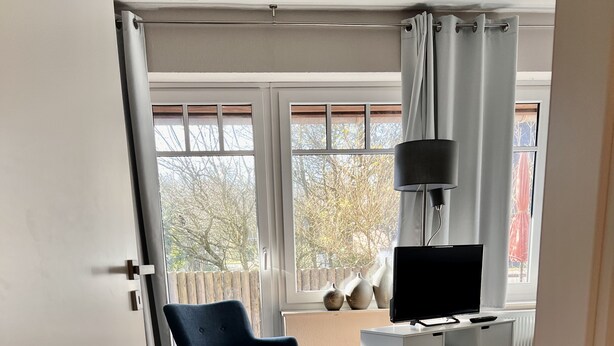 Una habitación con piso de madera, una silla azul, un escritorio blanco con televisor y una ventana con cortinas.