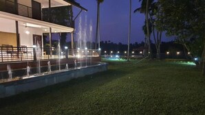 A nighttime view of a hotel with a water fountain feature in the foreground and a building with outdoor seating. - Lhasa Ayurveda and Wellness Resort (North Paravur)