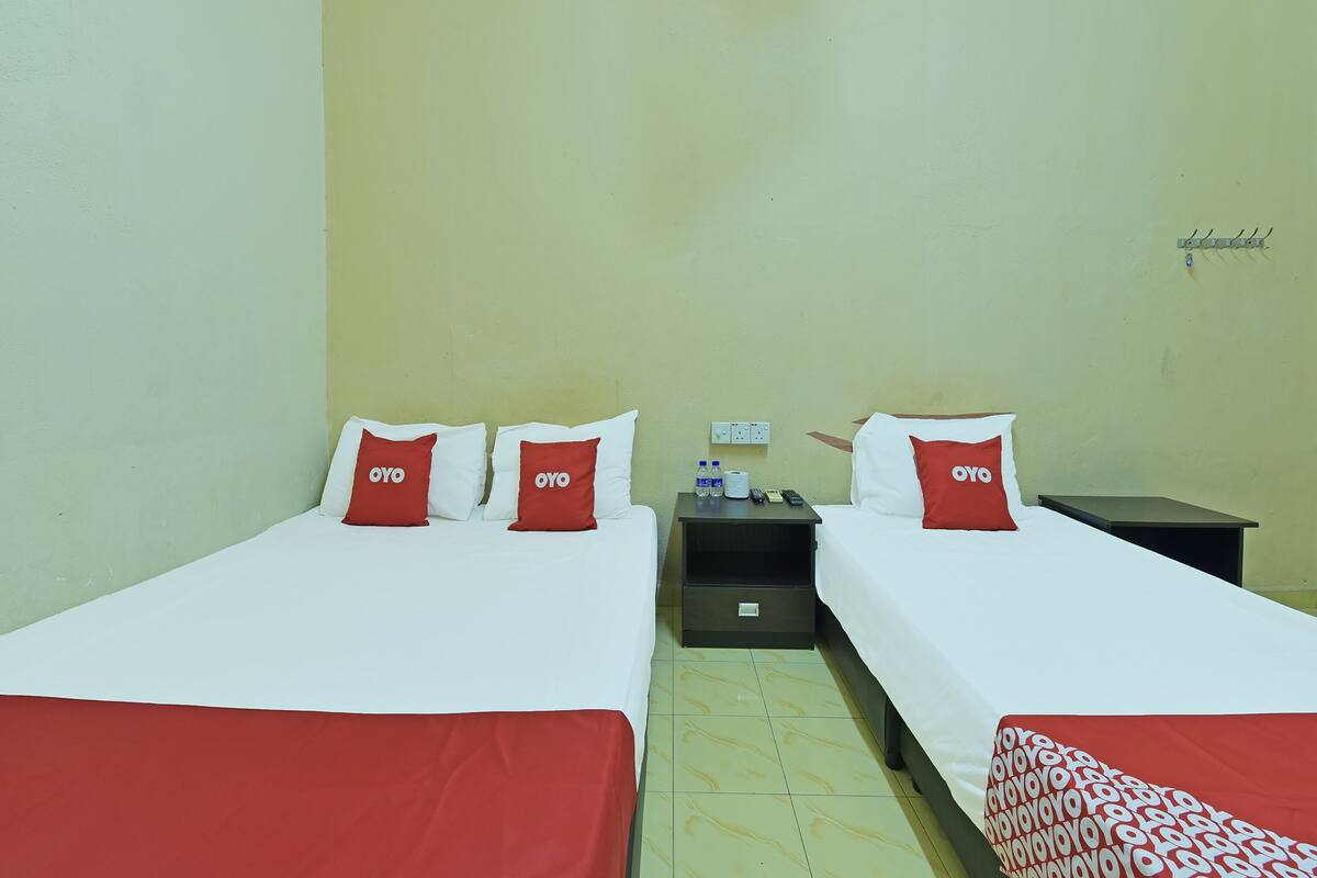 A hotel room with two beds, each with white bedding and red pillows labeled "OYO". There is a nightstand with a remote control, a bottle of water, and a tissue box. The room has a simple, functional design with a plain wall and a tiled floor.